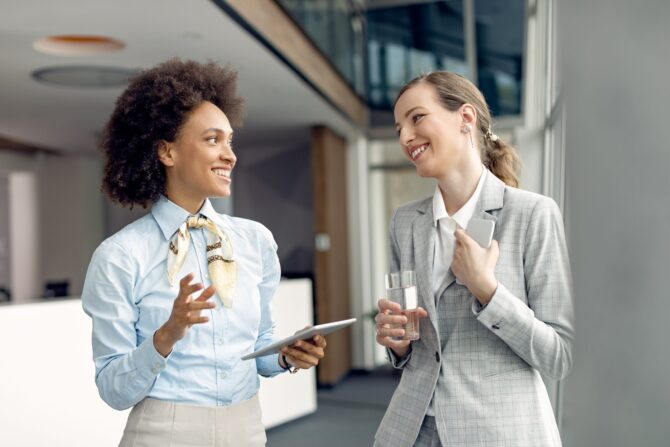 Two happy businesswomen communicating while standing in a hallwa