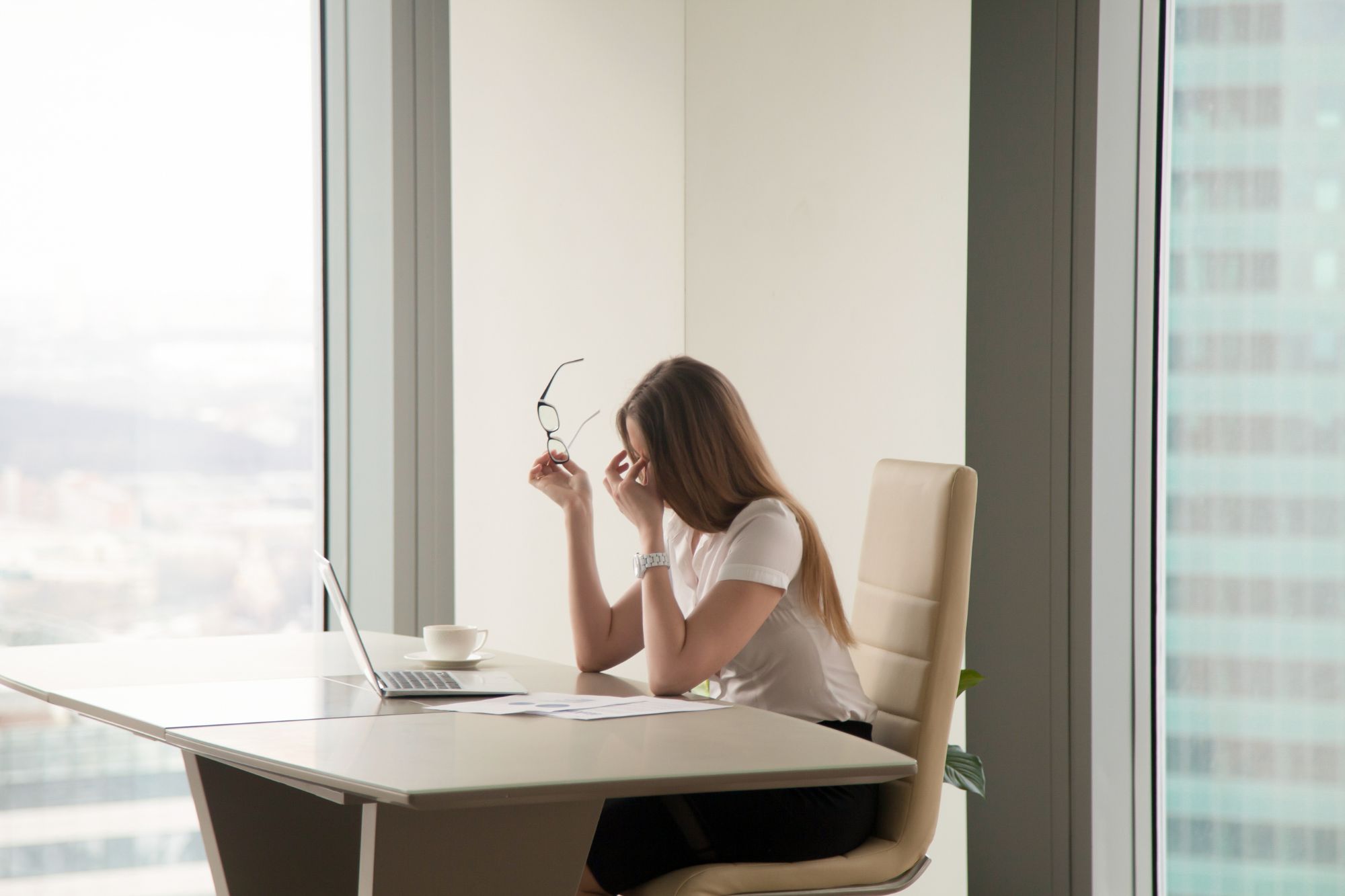 You Can’t See It, But You Can Feel It: Leadership Under Pressure Side profile of a stressed businesswoman sitting at her office desk by a large window, rubbing her eyes and holding her glasses in frustration with a laptop in front of her, illustrating a leader experiencing emotional exhaustion under pressure.