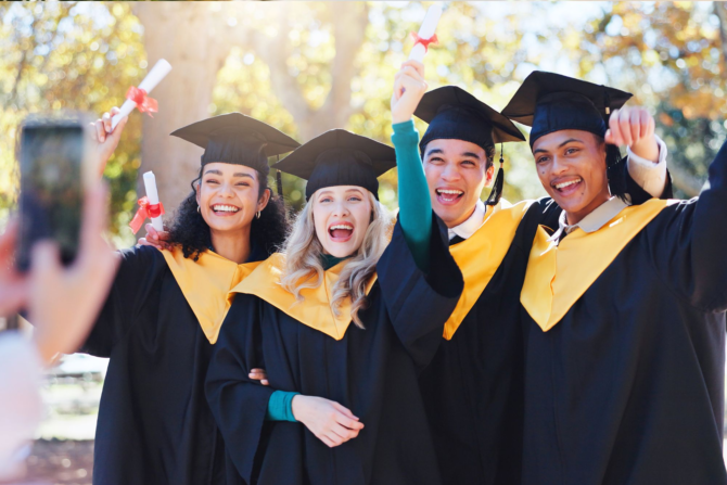 Becoming: Shift Focus From Performance to Human Growth | Return on Happiness A diverse group of four joyful university or high school graduates smiling, laughing, and holding up their diplomas during an outdoor celebration while someone takes their photo with a smartphone.