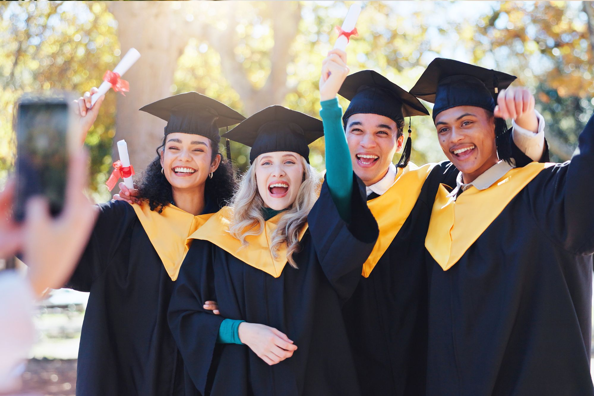 Becoming: Shift Focus From Performance to Human Growth | Return on Happiness A diverse group of four joyful university or high school graduates smiling, laughing, and holding up their diplomas during an outdoor celebration while someone takes their photo with a smartphone.