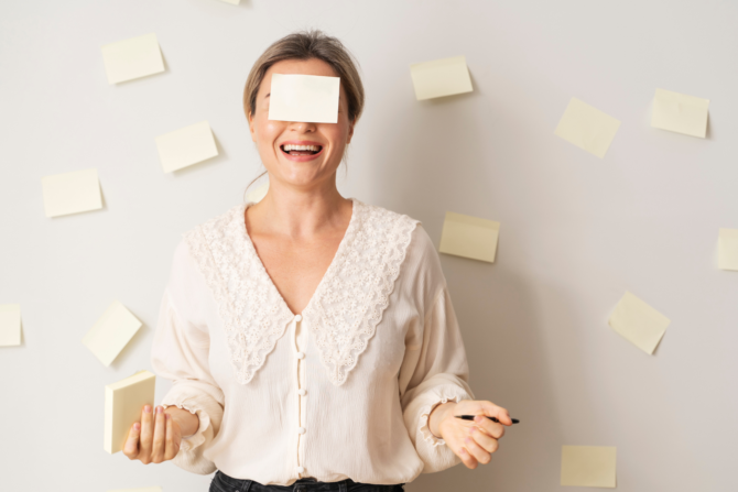 Leadership Patterns You Don’t See: Positive Leadership Awareness A laughing woman with a light yellow sticky note covering her eyes stands in front of a wall scattered with sticky notes, playfully representing invisible leadership patterns that leaders fail to see.