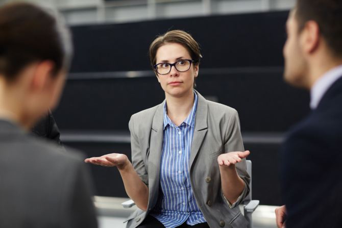 Is There One Question Every Leader Should Ask A female executive wearing glasses and a grey blazer holds her hands out in a questioning gesture during a meeting with two colleagues, illustrating a leader reflecting on the emotional climate they create.