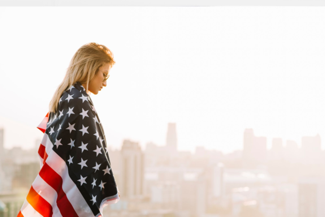 Today Is a Day of Remembrance: Leadership Reflection Side view of a blonde woman draped in an American flag, standing thoughtfully in contemplation with a blurred city skyline background during sunset.
