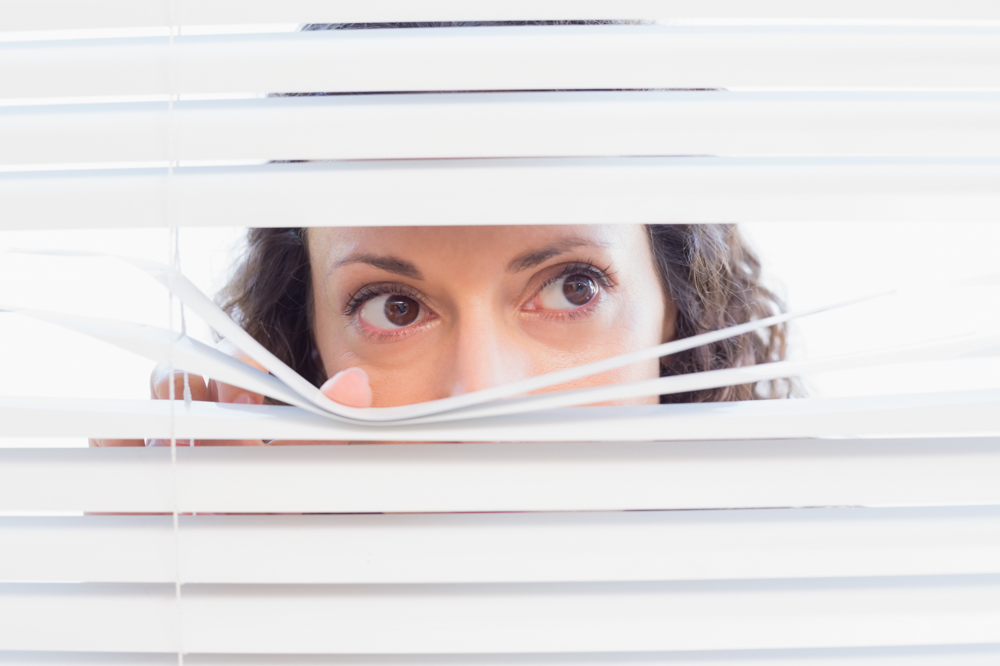 What Leadership Patterns Are Still Hiding Close-up of a woman peering cautiously through white window blinds, representing a leader uncovering hidden leadership patterns that affect employee engagement